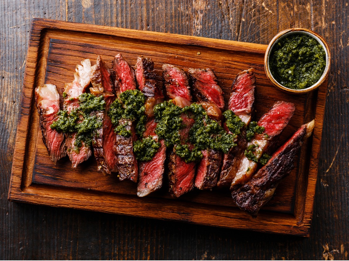 plano zenital carne troceada a la parrilla sobre mesa de madera y salsa chimichurri